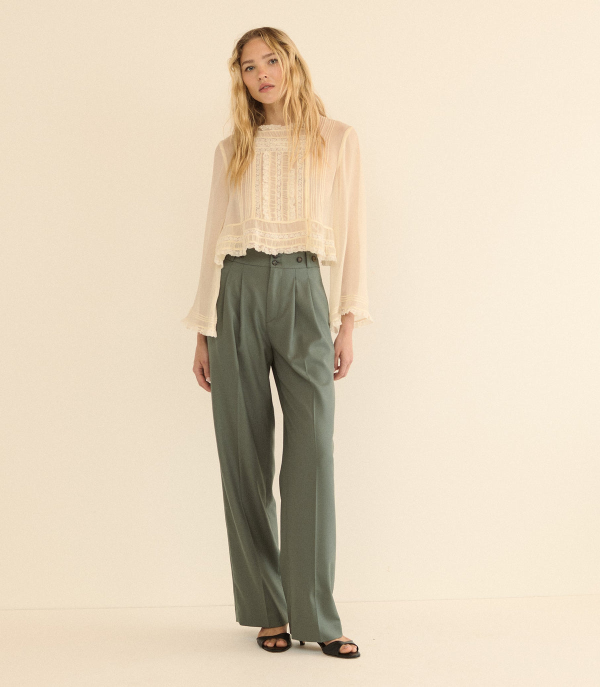 A woman stands against a plain warm cream studio background wearing a sheer long-sleeve cream blouse with a lace front and high-waisted olive green wide-leg trousers.