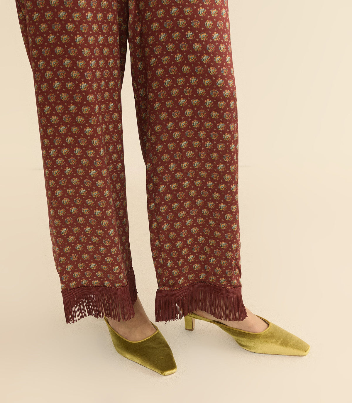 A close-up of a person's legs wearing rust-brown wide-leg trousers with a small floral pattern and fringe hems, paired with gold pointed-toe heels on a light beige background.